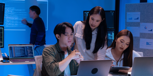 A group of young Asian technology professionals collaborating in a modern office with blue ambient lighting, looking closely at laptop screens and monitor displays working alongside AI
