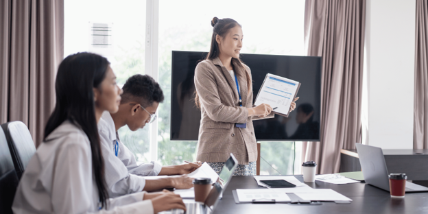 An Asian female business professional in a beige blazer standing up and presenting a printed report or chart to colleagues sitting around a conference room table.